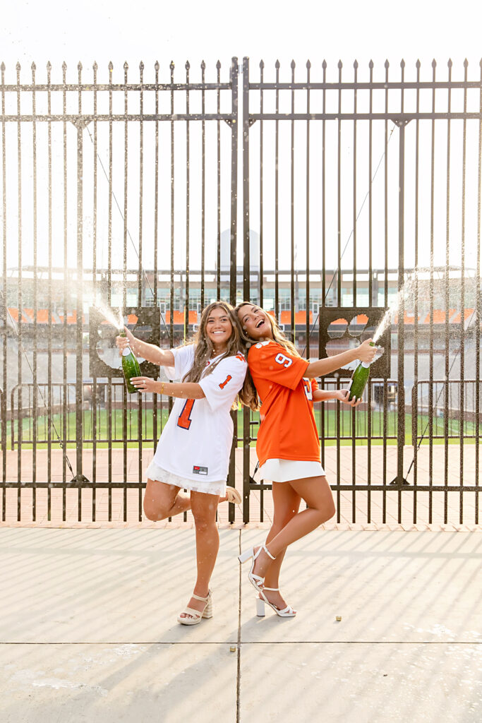 Two Clemson University seniors & graduates popping bottles of champagne outside of memorial stadium in front of the Clemson paw gates wearing orange and white Clemson football jersey.s